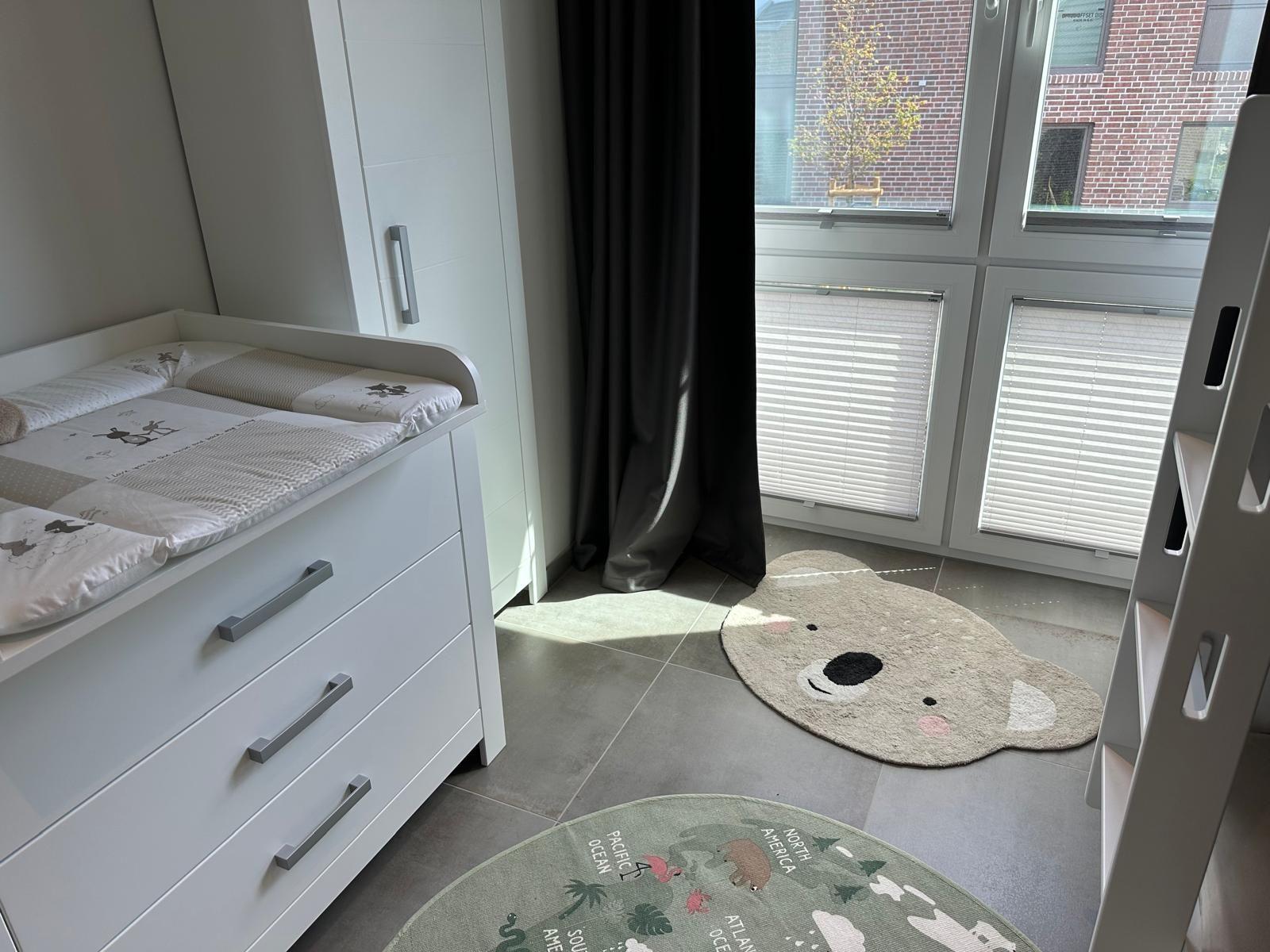 Kinderzimmer mit Wickelkommode, Bären-Teppich und Fenster mit Jalousien.