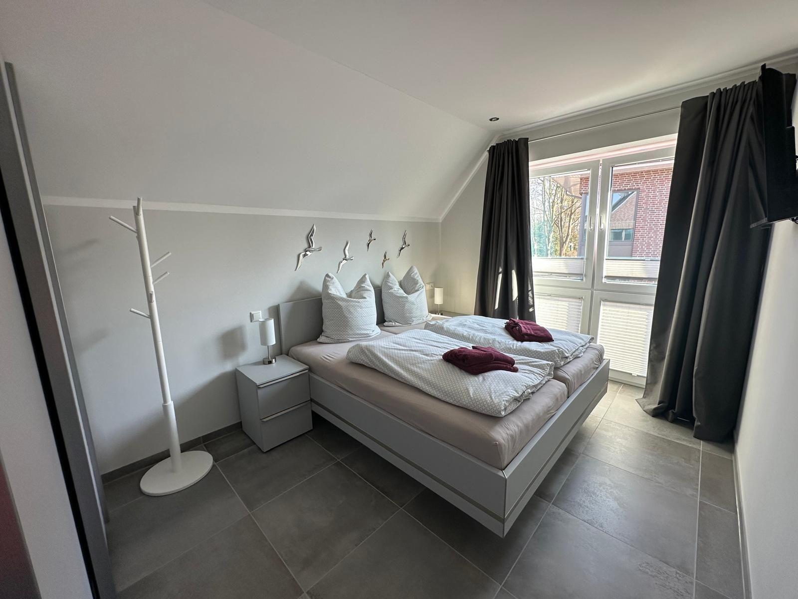 Schlafzimmer mit Doppelbett, Nachttisch und Kleiderständer. Fenster mit Vorhängen.