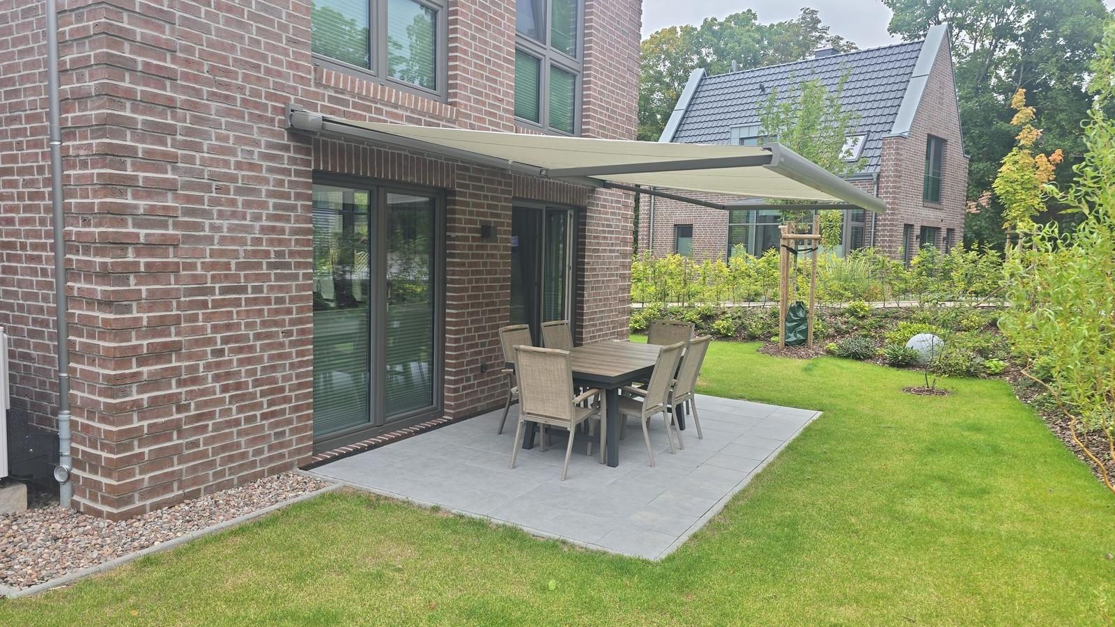 Terrasse mit Tisch und Stühlen unter Markise. Grünfläche und Gartenhaus im Hintergrund.