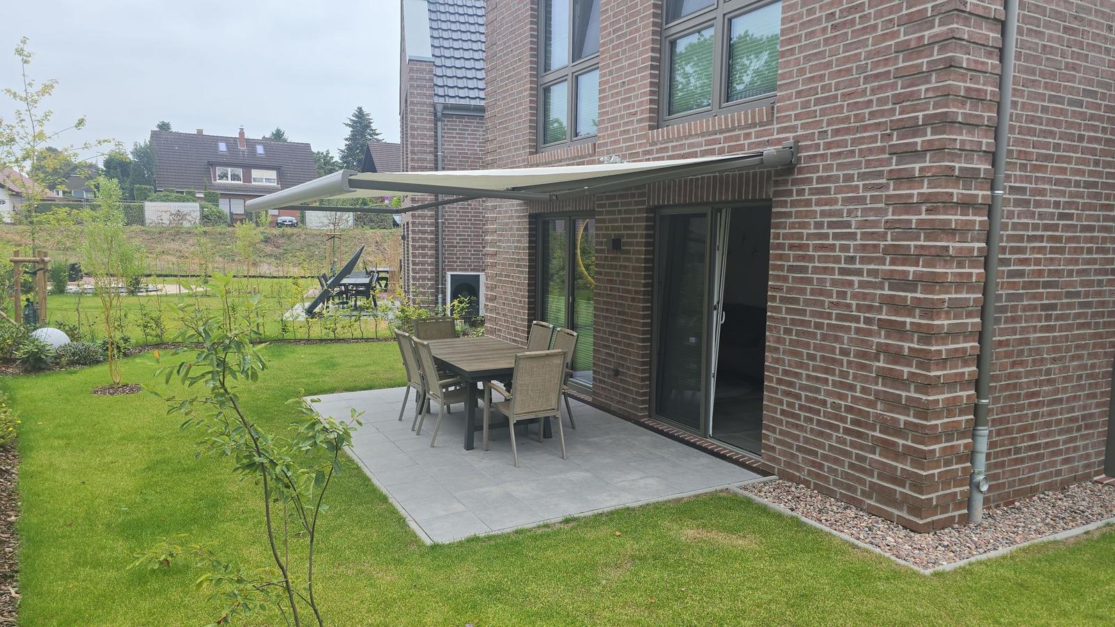 Terrasse mit Tisch und Stühlen unter Markise. Grünflächen und Gartenbereich.