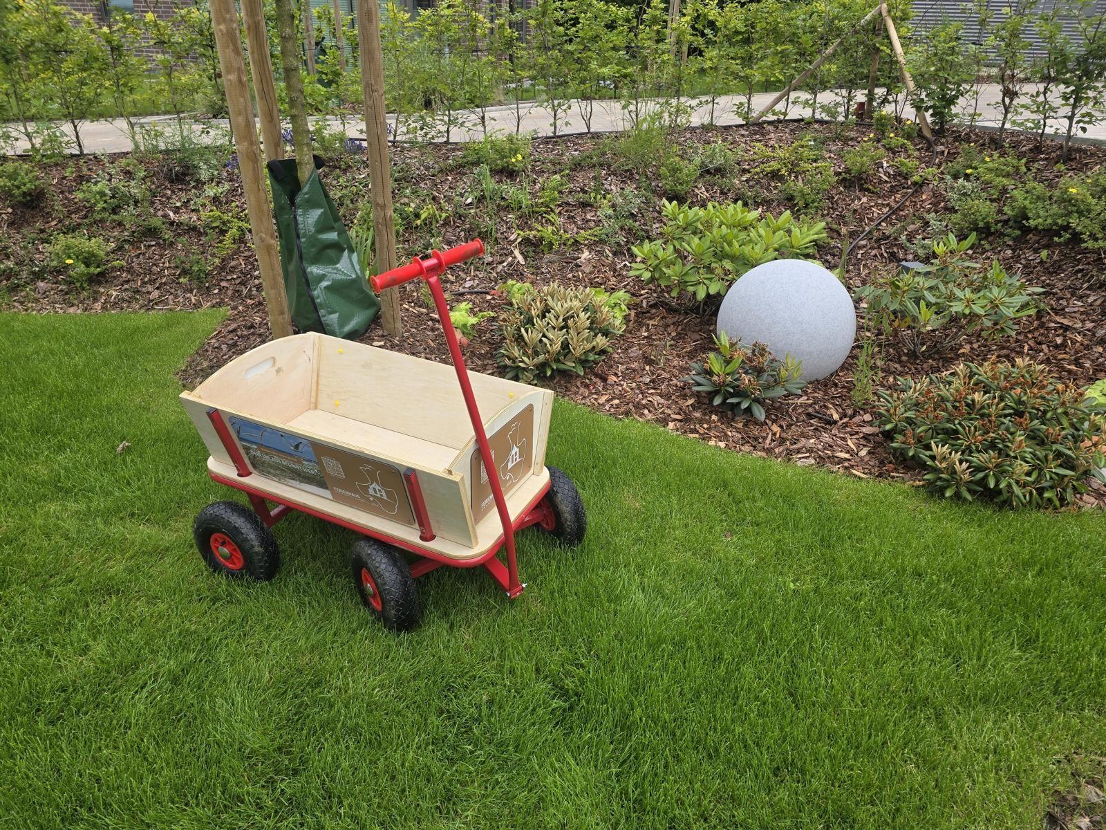 Ein roter Kinderwagen steht auf dem Rasen neben einem Beet mit Pflanzen und einem Steinball.