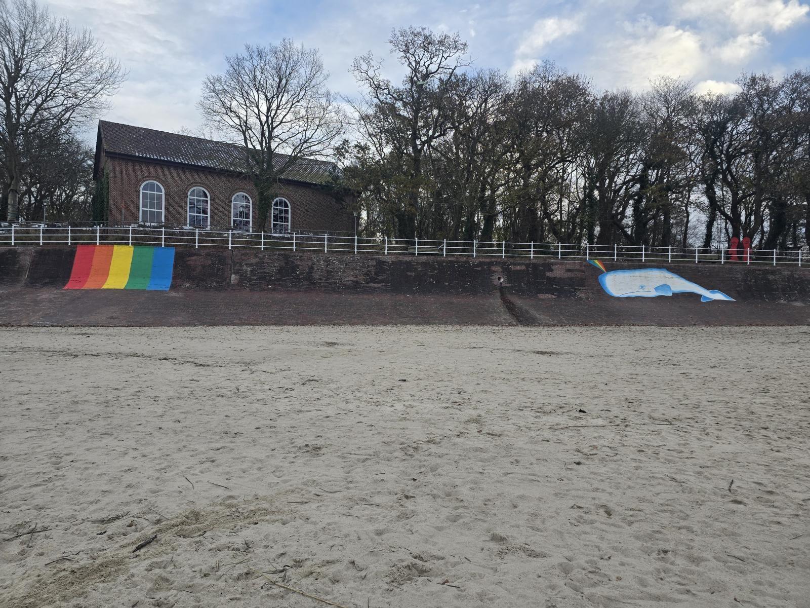 Strand mit gemalter Wal und Regenbogenwand vor einer Backsteinhütte.