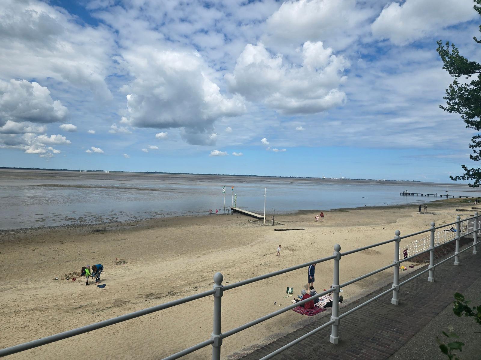 Strand mit Watt, Menschen, Steg und blauem Himmel mit Wolken.