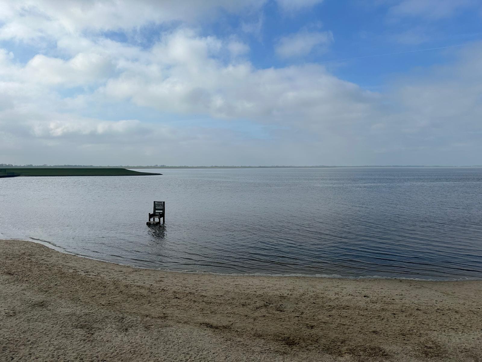 Eine Stuhl am Strand im Wasser, Blick auf ruhiges Gewässer und Himmel mit Wolken.