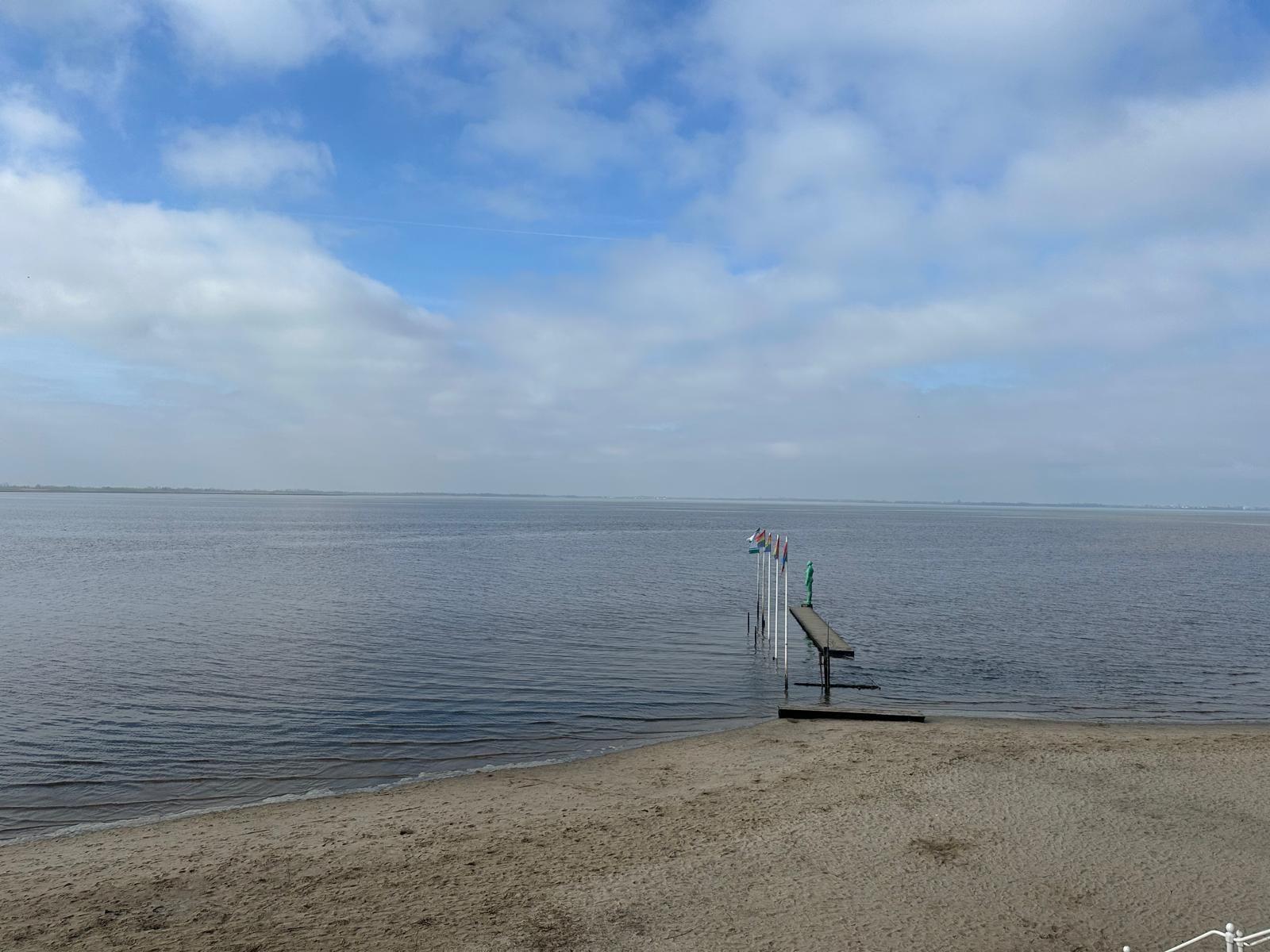Strand mit Steg und Flaggen am Wasser. Blick auf ruhiges Gewässer unter bewölktem Himmel.