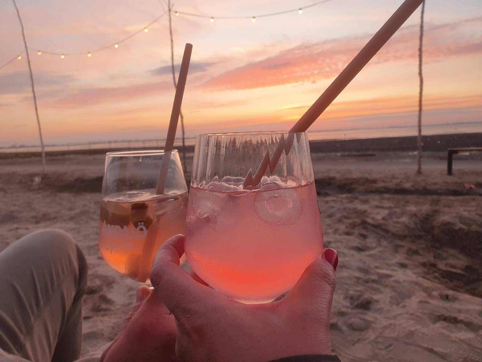 Zwei Gläser mit Getränken auf dem Strand bei Sonnenuntergang mit Lichterkette im Hintergrund.
