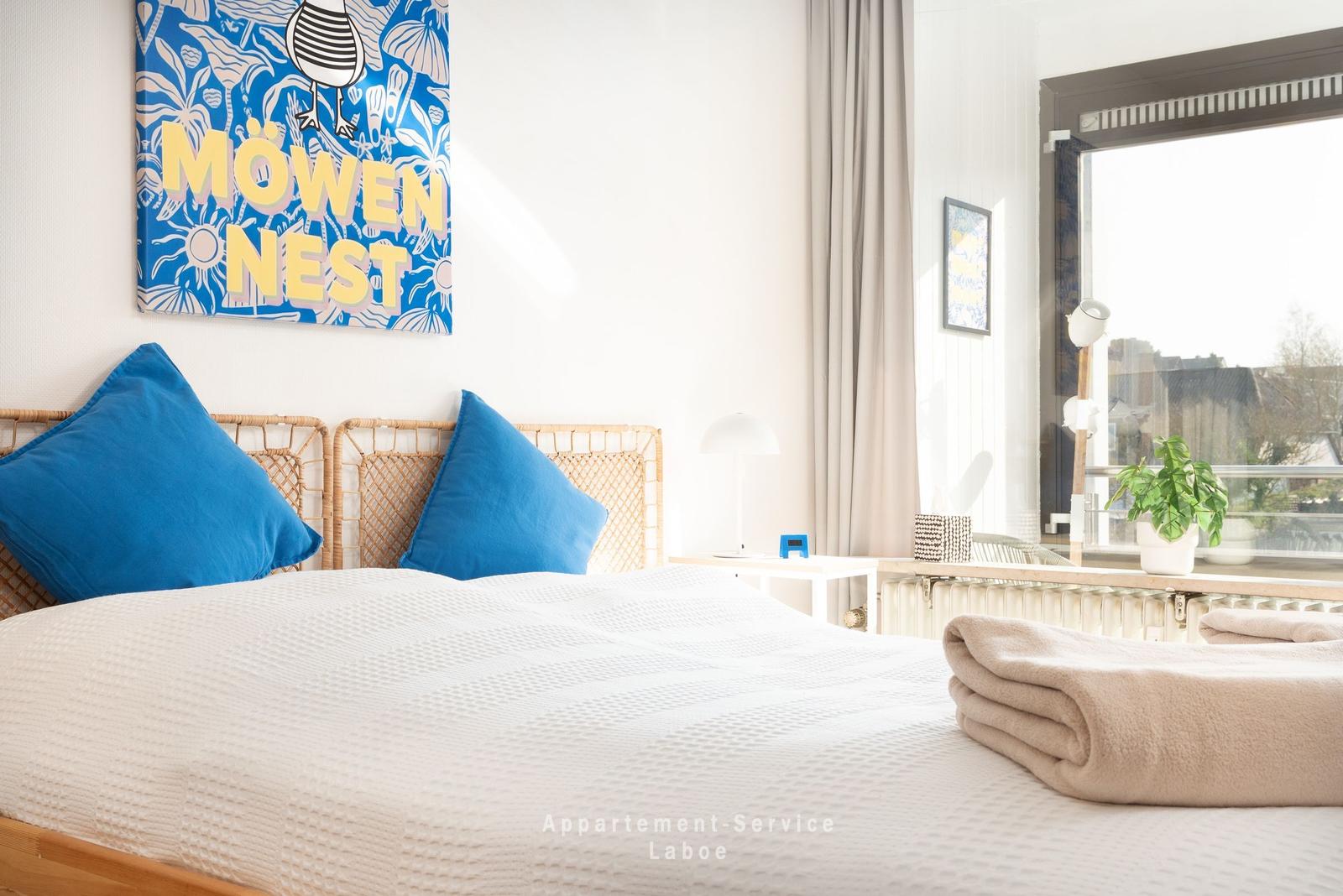 Ein Schlafzimmer mit Bett, Kissen und Wandbild. Fenster mit Blick nach draußen.
