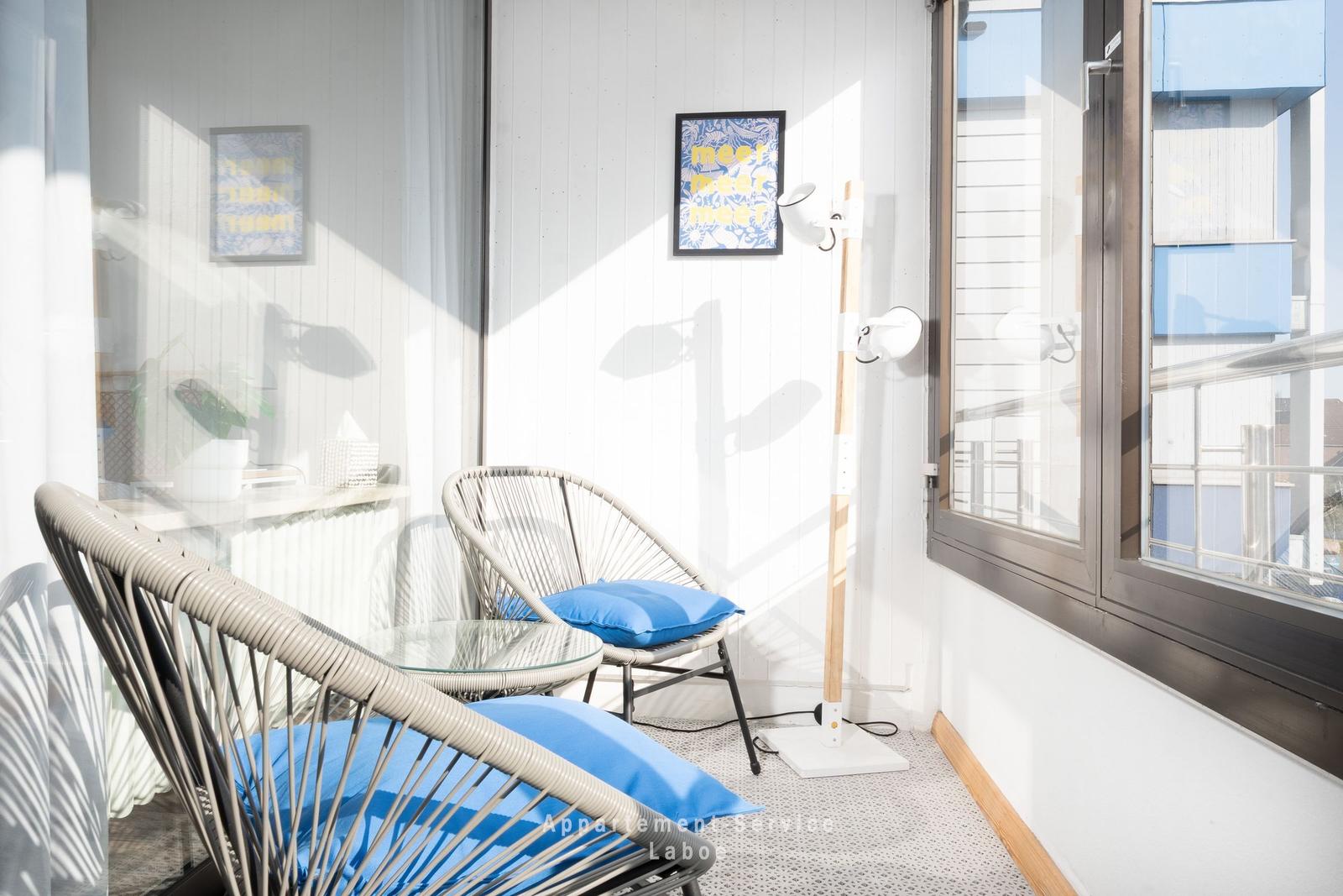 Terrasse mit zwei Stühlen, Tisch und Stehlampe. Wandbild und Fenster sichtbar.