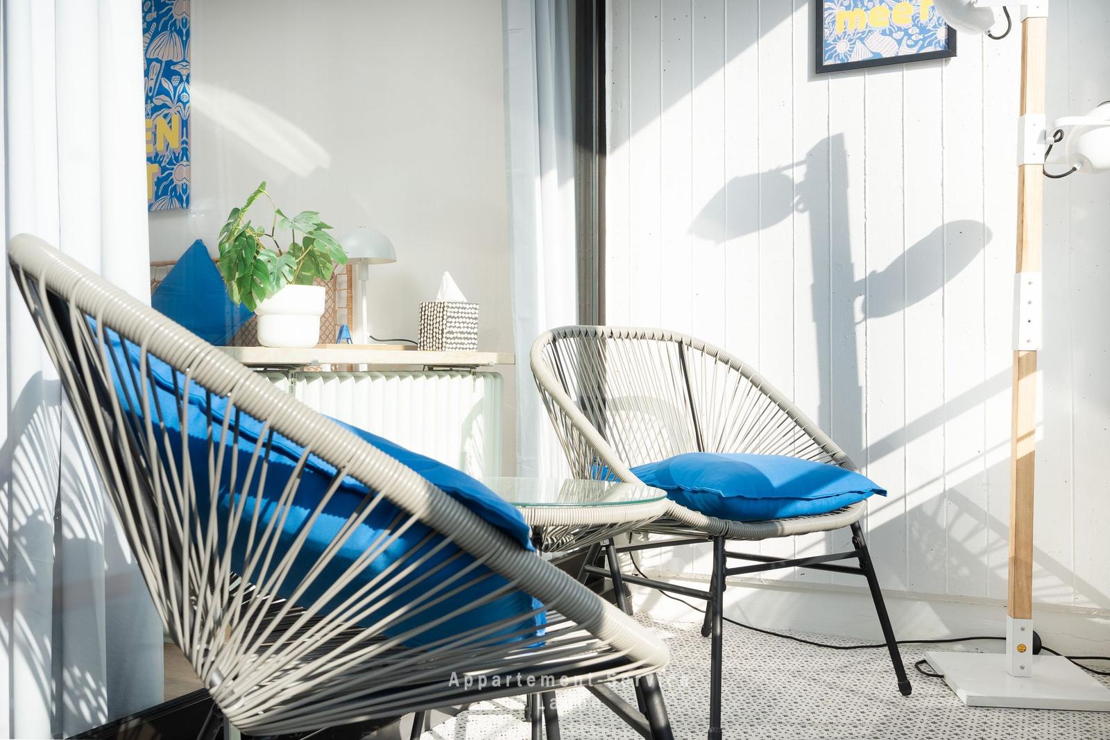 Zwei Stühle mit blauen Kissen und Glas-Tisch auf Balkon mit weißen Wänden und Pflanze.