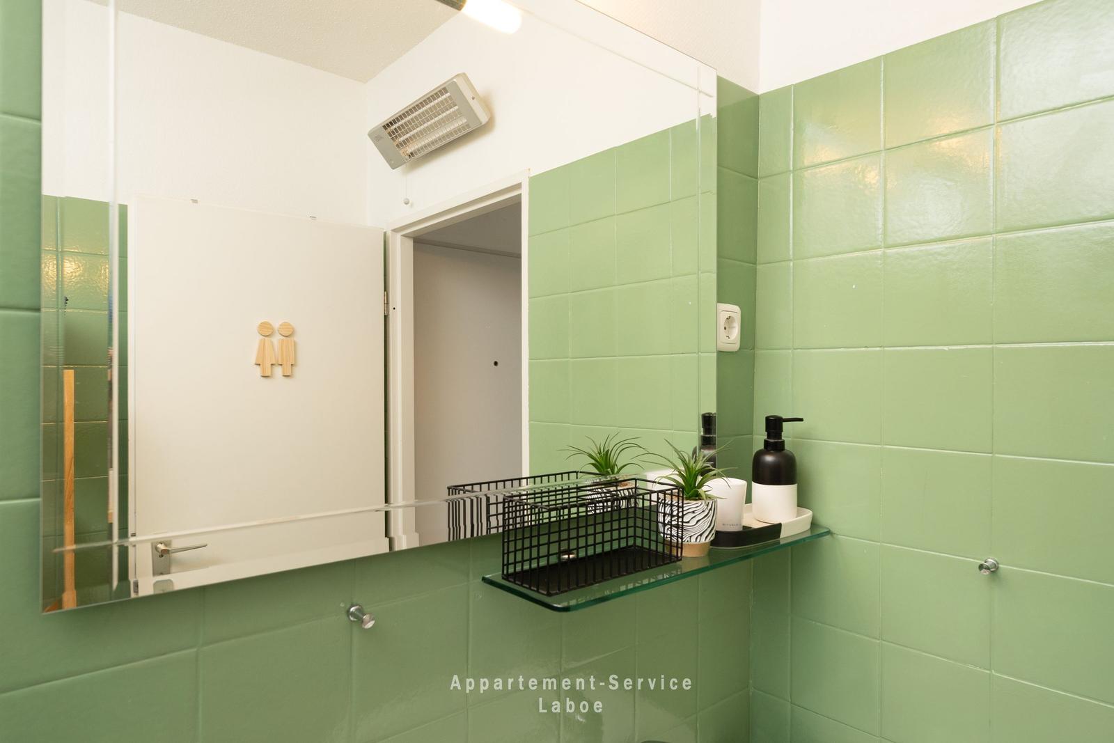 Badezimmer mit grünen Fliesen, Spiegelschrank und Glasregal mit Pflanzen und Duschbereich.
