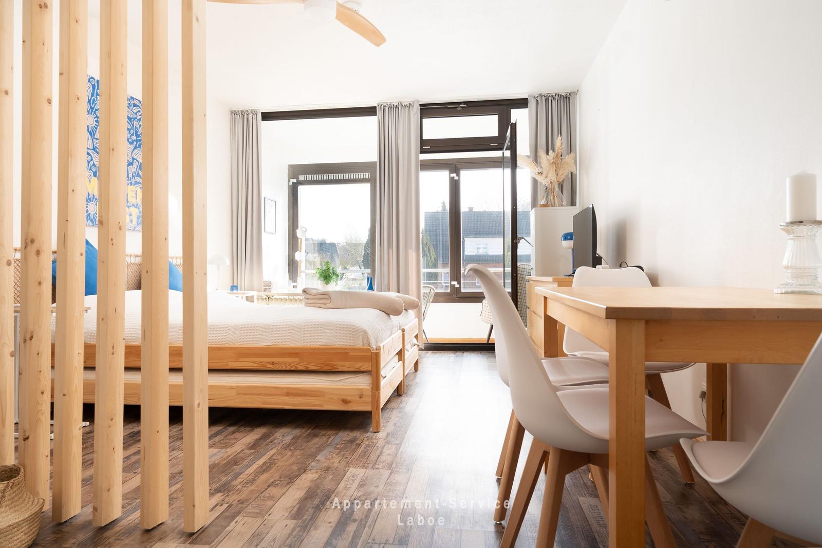 Ein Schlafzimmer mit Holzbett und Trennwand. Es gibt einen Esstisch mit Stühlen und einen Balkon.