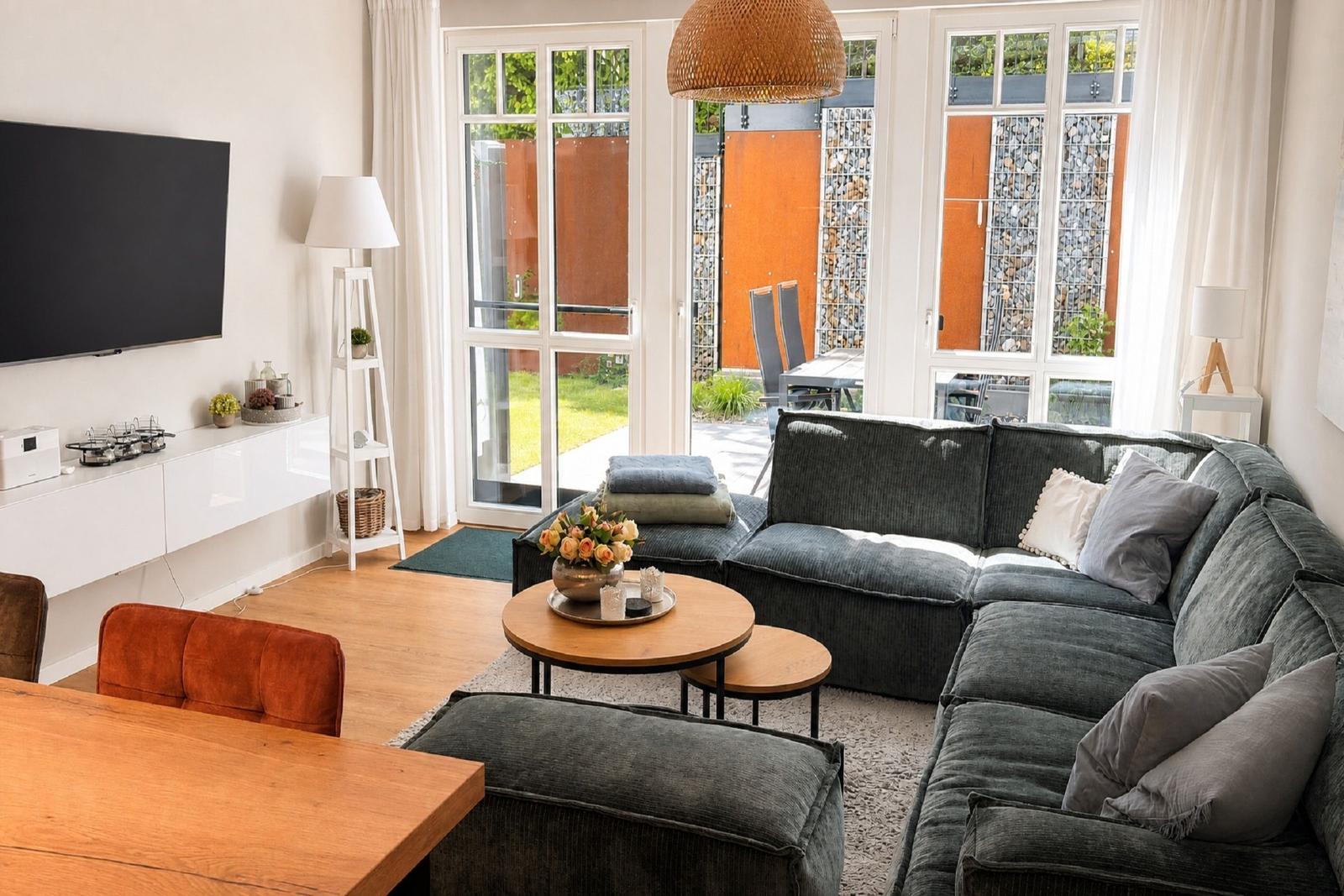 Wohnzimmer mit Sofa, Couchtisch und Fernseher. Durchgang zur Terrasse mit Garten.