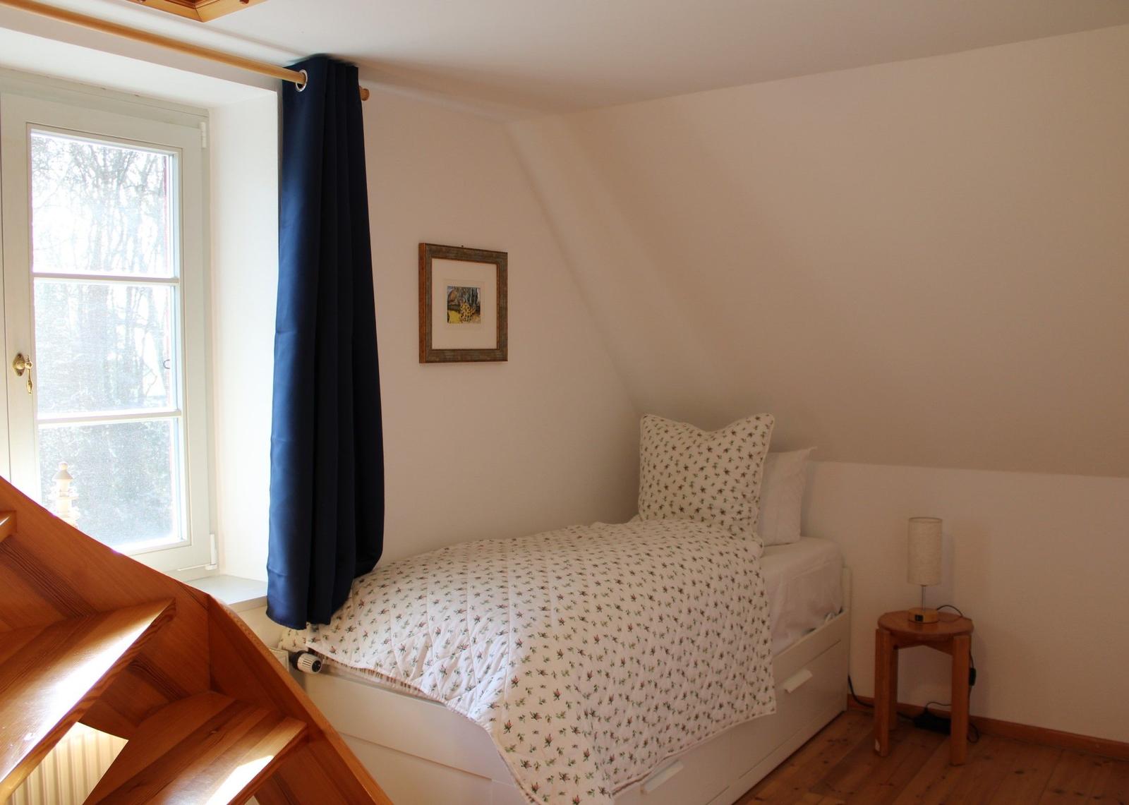 Ein Schlafzimmer mit Dachschrägen, Bett, Nachttisch und Fenster mit Blick auf Wald.