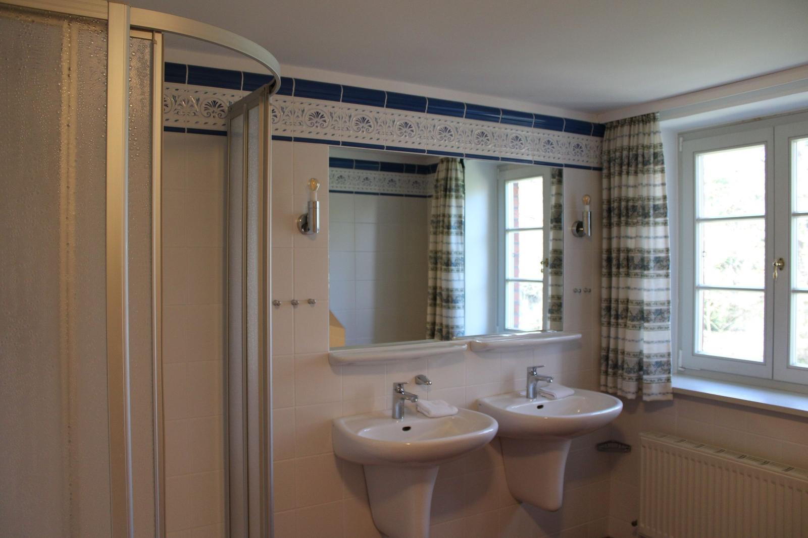 Badezimmer mit Dusche, zwei Waschbecken und Fenster mit Vorhang.