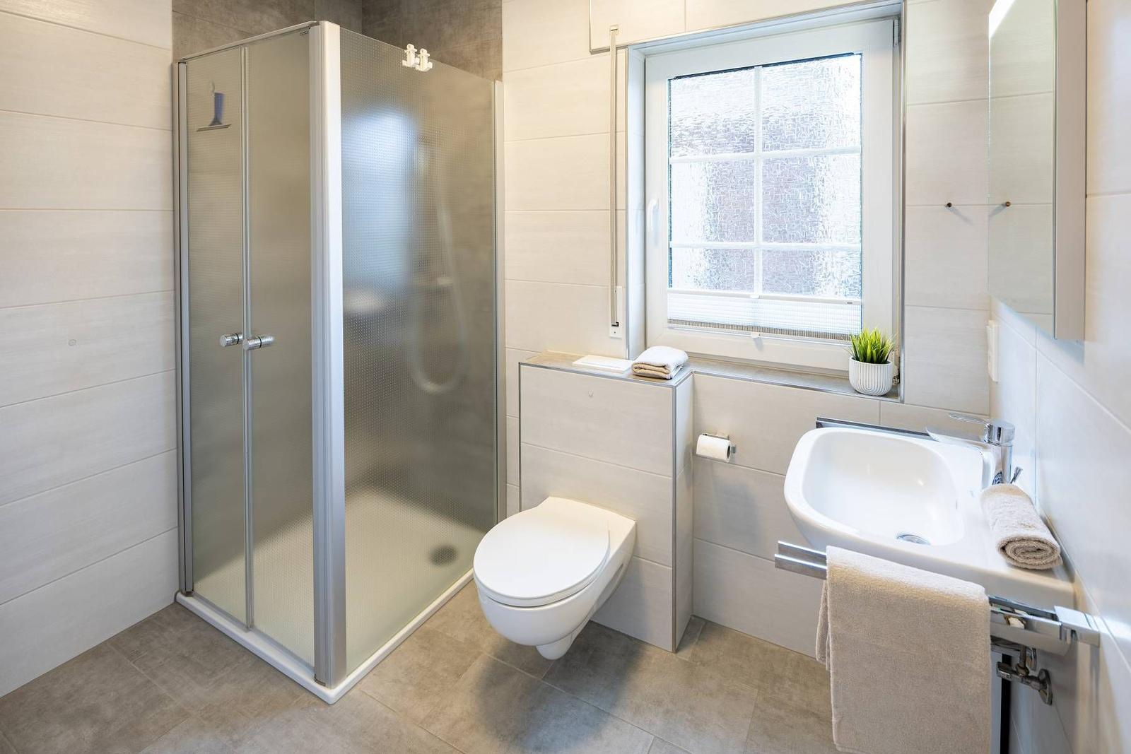 Badezimmer mit Dusche, Toilette und Waschbecken. Fenster mit Pflanze.