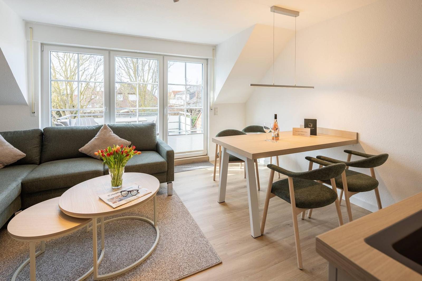 Wohn- und Essbereich mit Sofa, Tisch und Stühlen. Große Fenster mit Blick auf Balkon.