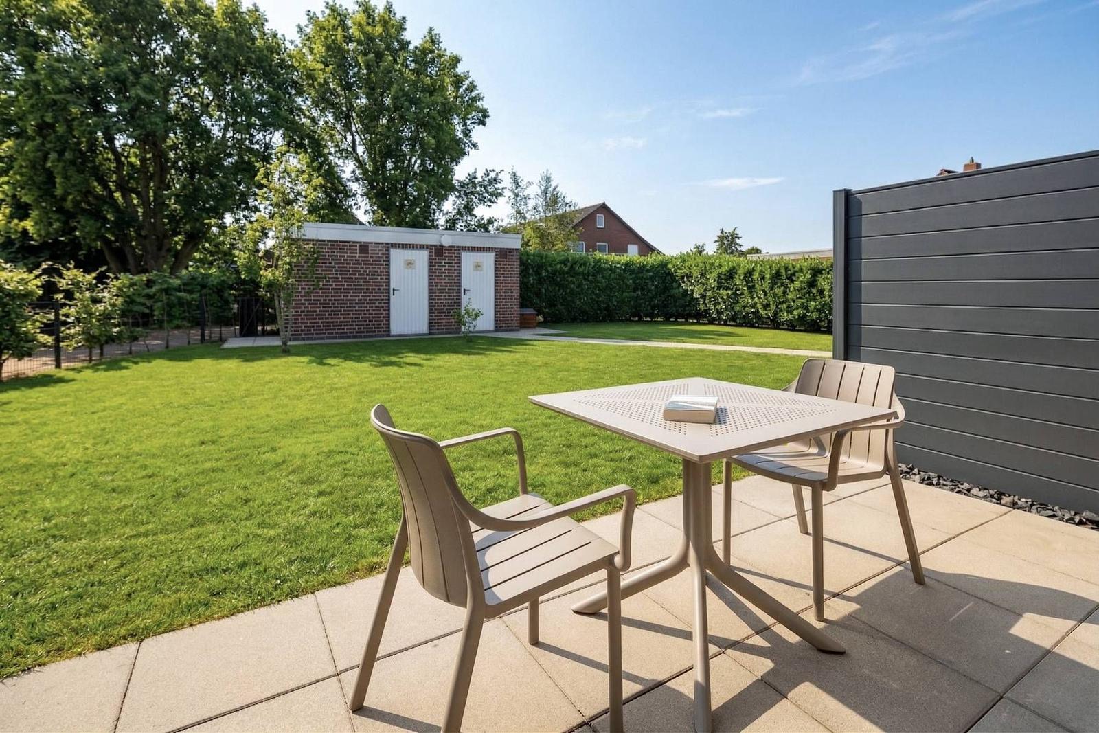 Terrasse mit Tisch und zwei Stühlen. Hintergrund: Rasenfläche, Hecke und Gebäude.