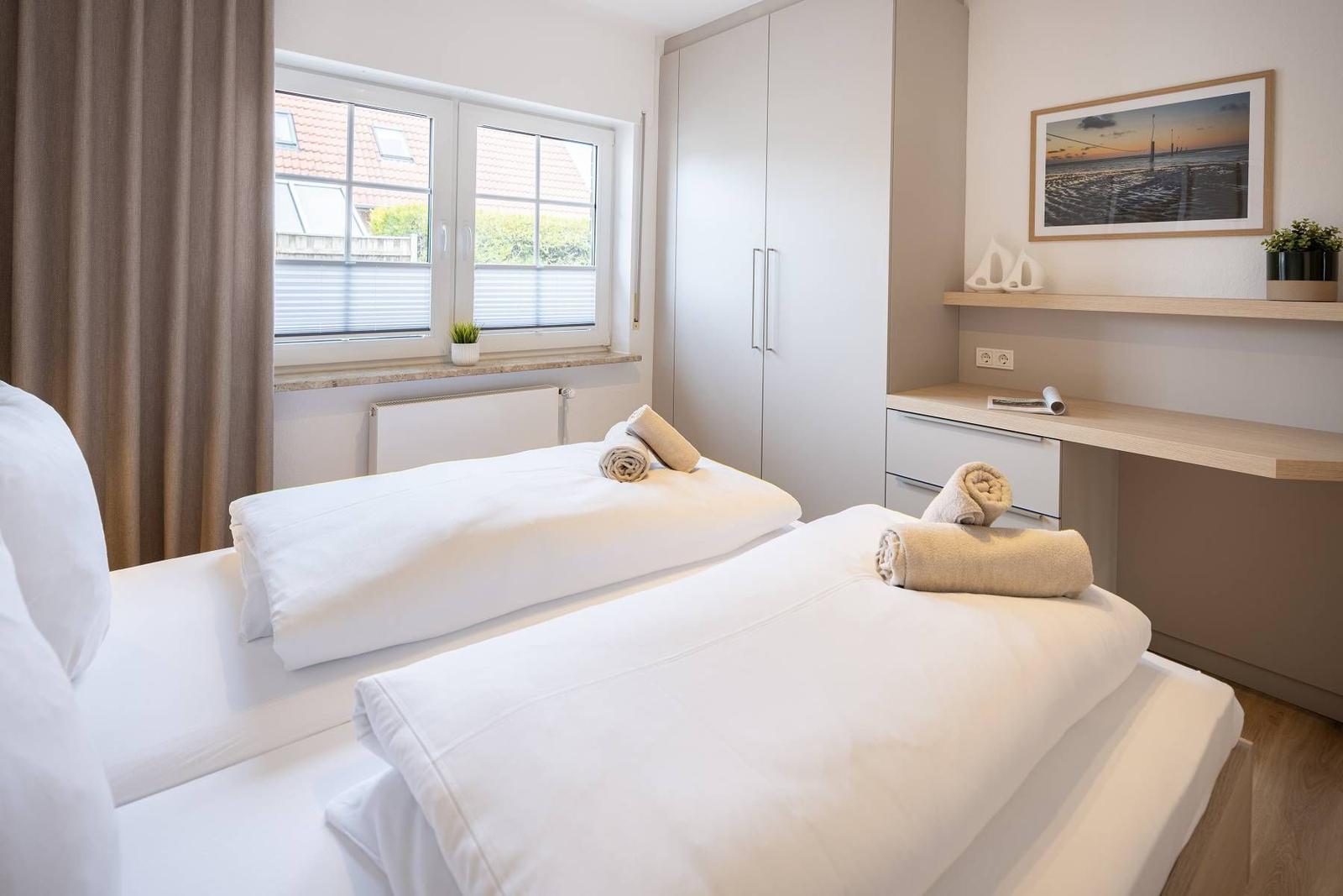 Zwei Betten mit weißen Bettwäsche, Fenster mit Blick auf Dächer, Schrank und Schreibtisch mit Bild.