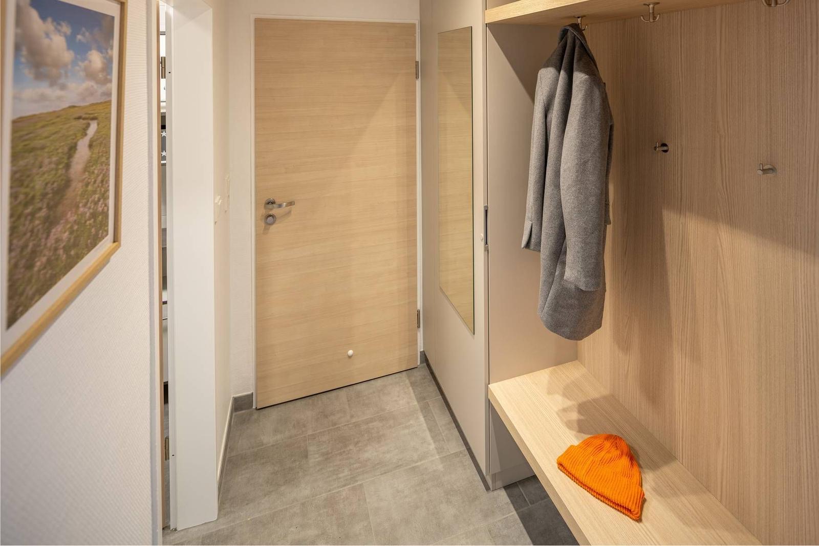 Flur mit Kleiderschrank, grauem Mantel und orangefarbener Mütze. Holztür und Wandbild sichtbar.