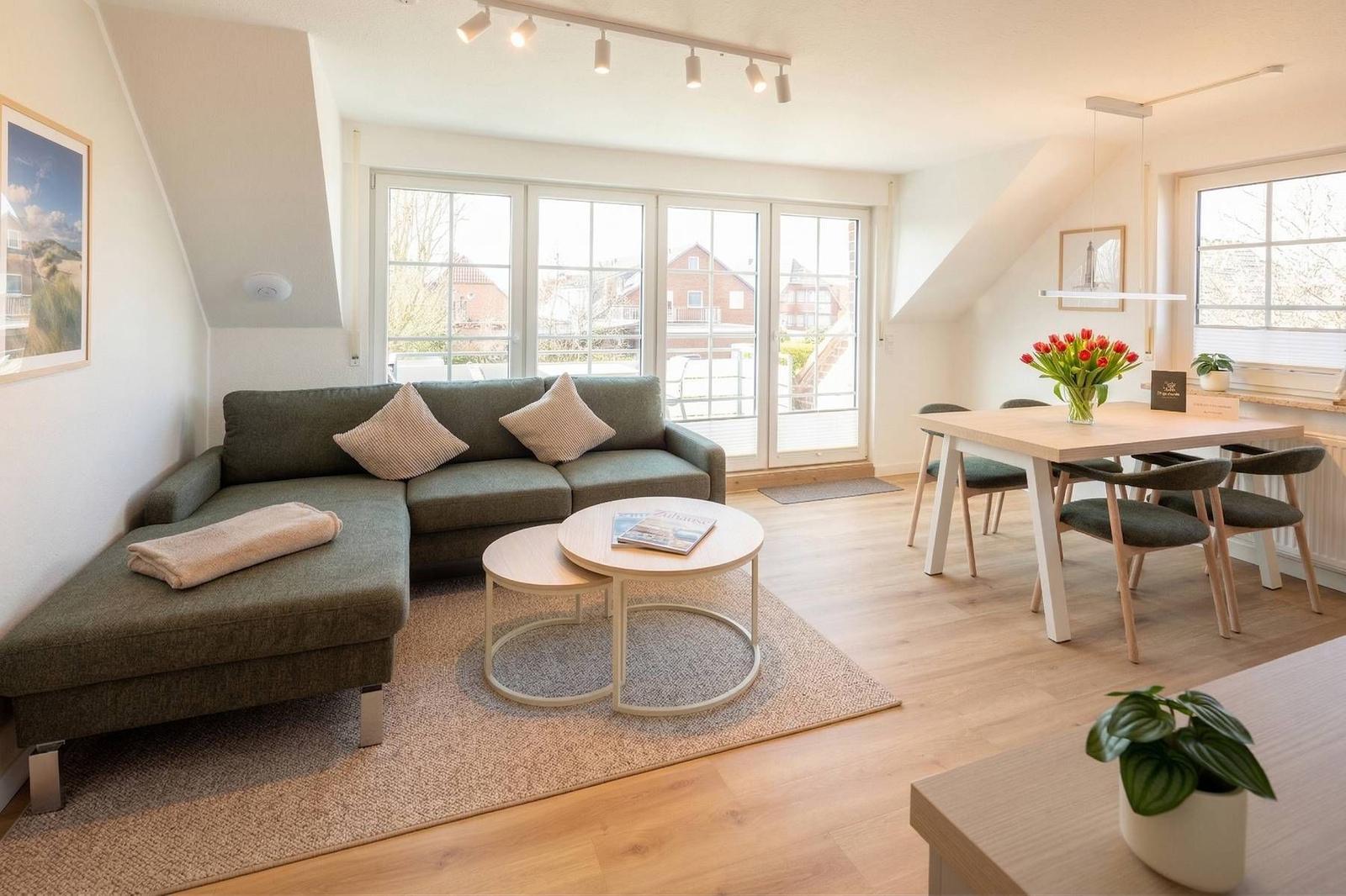 Wohn- und Essbereich mit Sofa, Tisch und Stühlen. Holzboden und große Fenster.