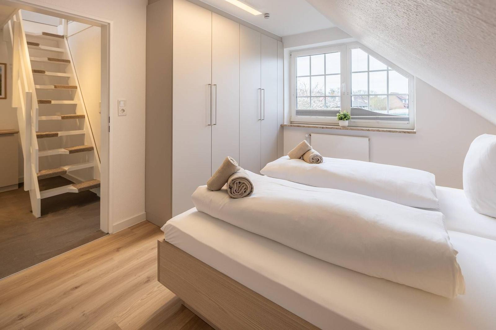 Zimmer mit Doppelbett, Kleiderschrank und Dachschrägenfenster. Treppenhaus im Hintergrund.