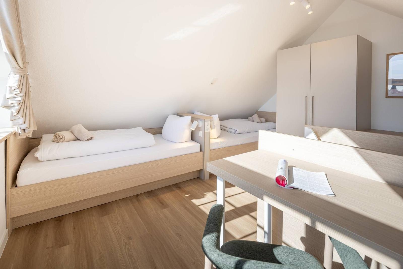 Zwei Betten, Schreibtisch und Kleiderschrank in einem Dachzimmer mit Holzboden und Fenster.
