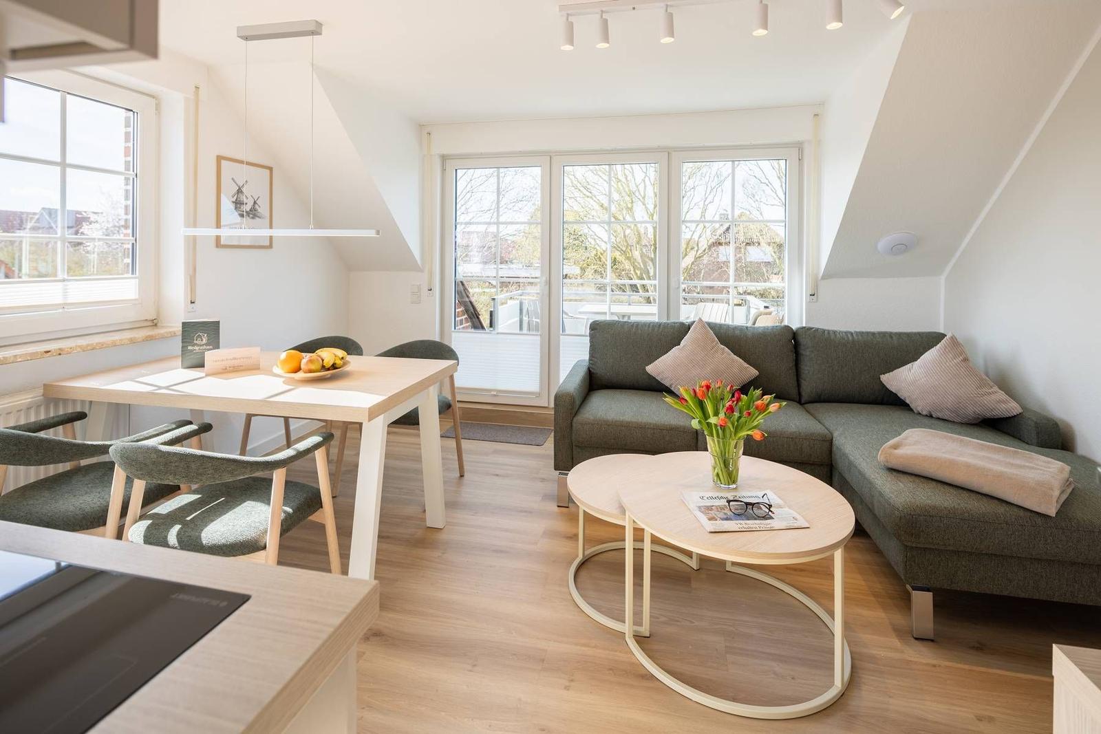 Wohn- und Essbereich mit Sofa, Tisch und Fenstern. Holzboden und helle Wände.