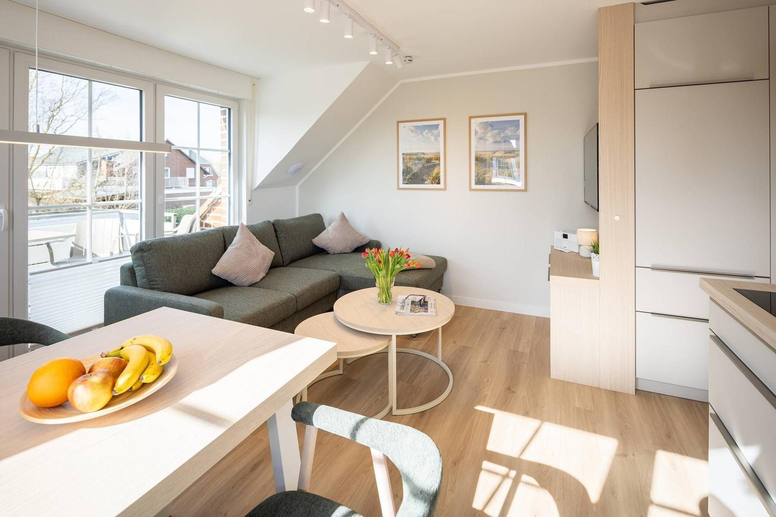 Wohnbereich mit Sofa, Esstisch und Küchenzeile. Große Fenster und Holzboden.