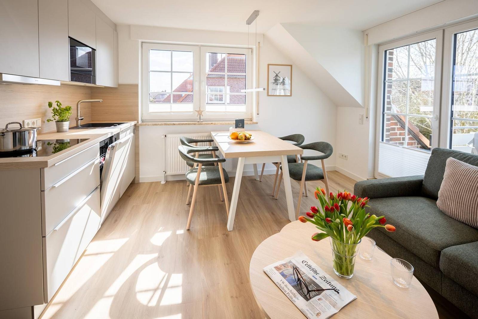 Küche, Essbereich und Wohnbereich mit Sofa, Tisch und Stühlen. Fenster mit Blick auf Dächer.