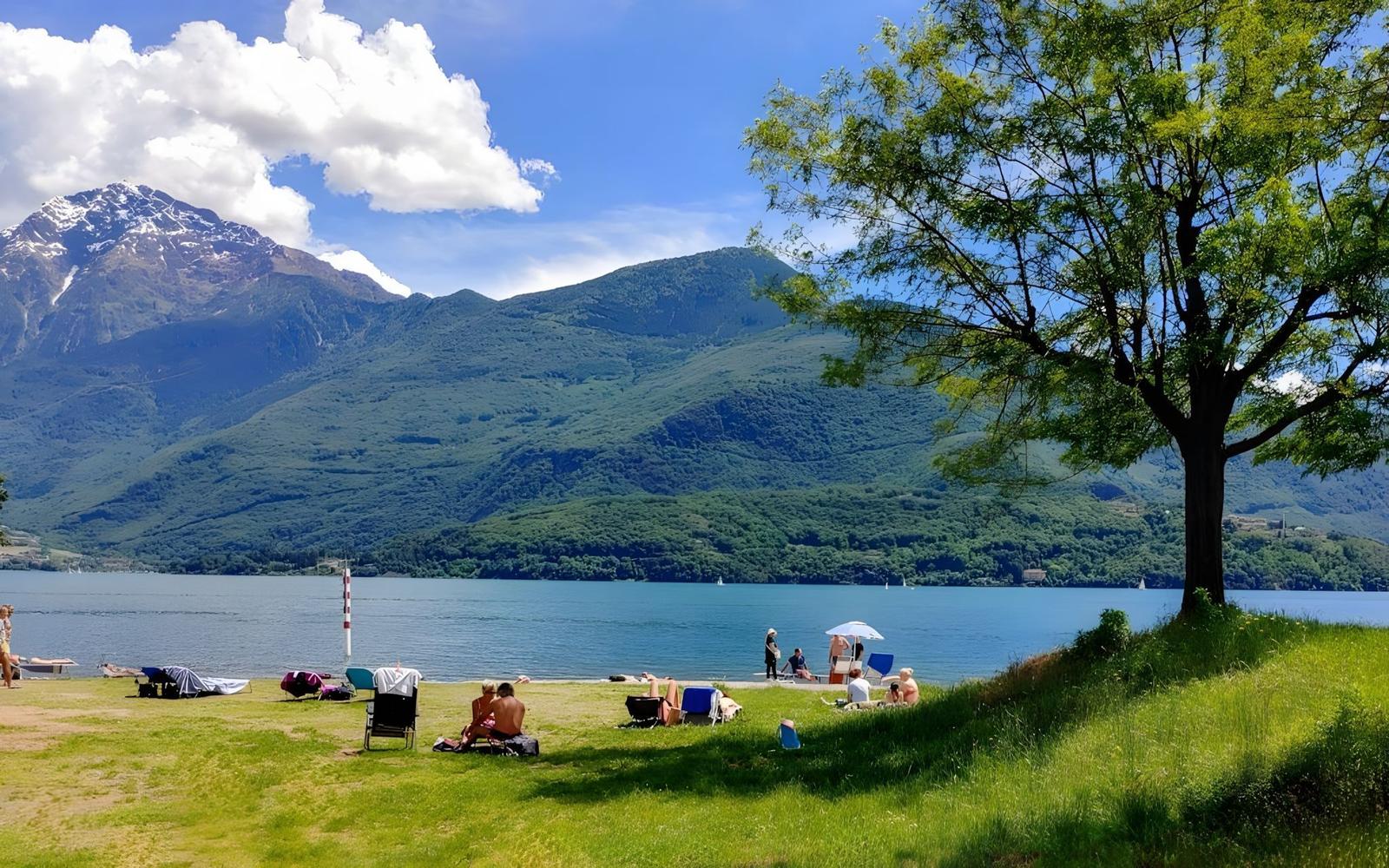 Grüner Strand mit Bäumen, Menschen und einem See vor Bergen.