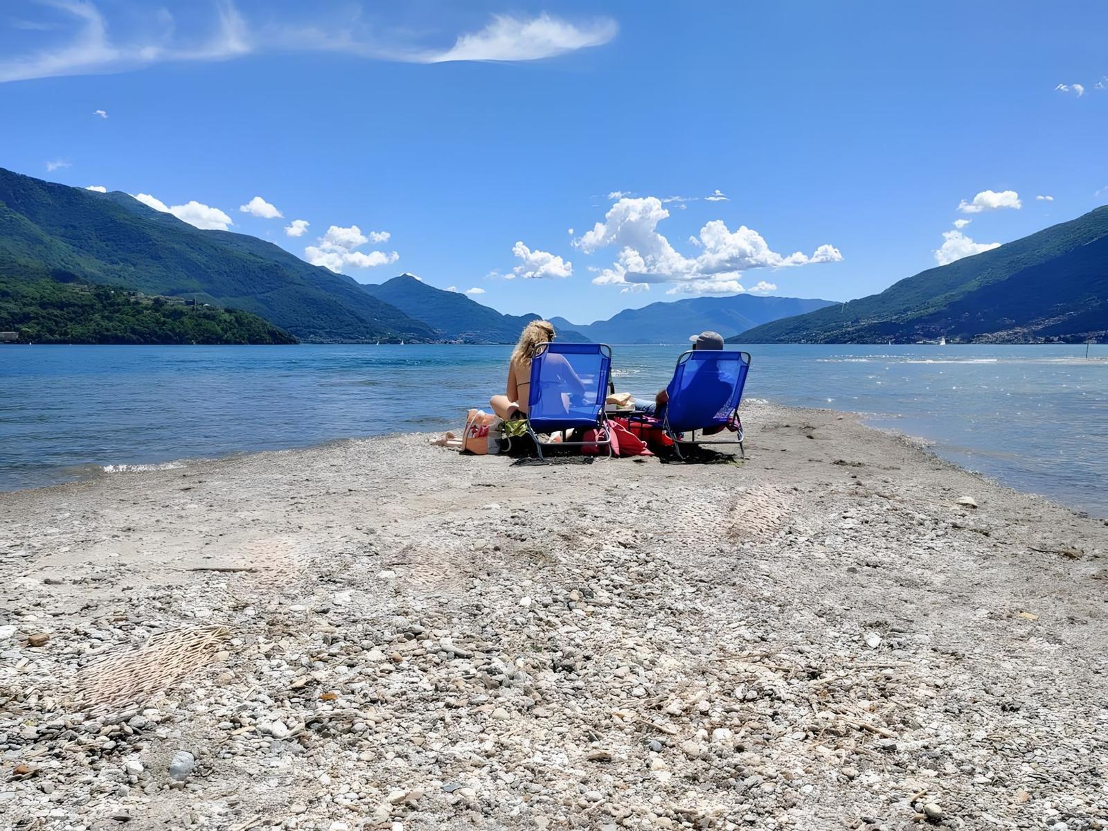Zwei Personen sitzen in blauen Liegestühlen am steinigen Ufer eines Sees mit Bergen im Hintergrund.