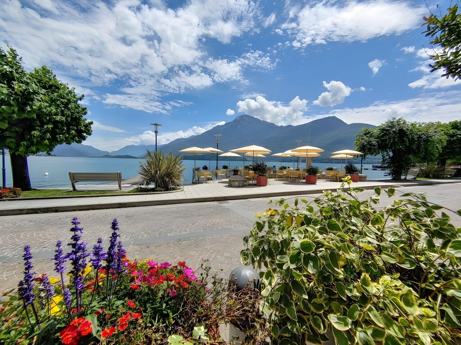 Terrasse mit Sonnenschirmen am See, umgeben von Blumen und Bergen.