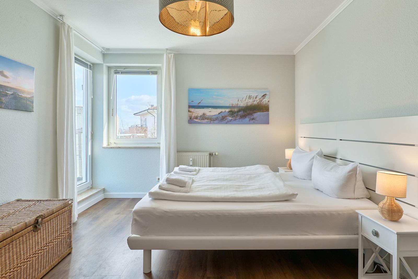Schlafzimmer mit weißem Bett, Holzboden und Fenster mit Blick auf Außenbereich.