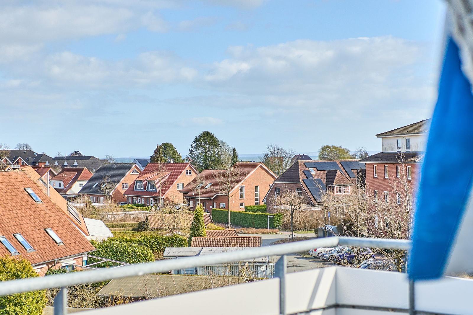 Blick von Balkon auf Wohnhäuser mit Dächern und Bäumen unter blauem Himmel.
