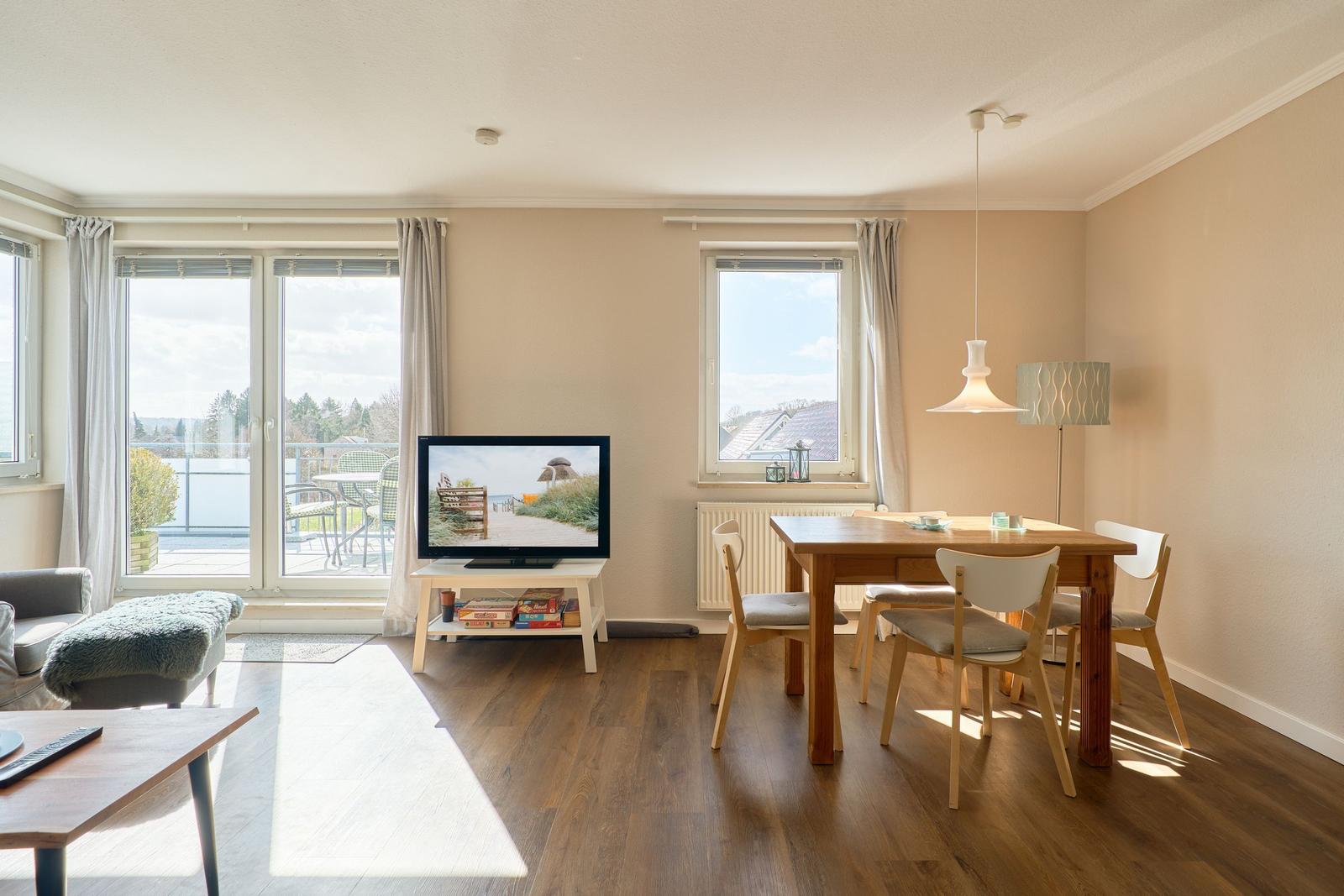 Wohn- und Essbereich mit Holzboden, TV, Tisch und Stühlen sowie Balkonansicht.