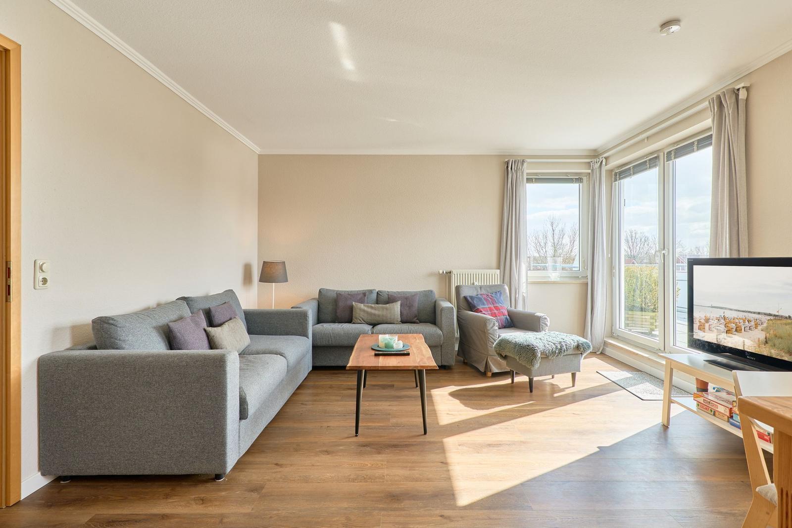 Wohnzimmer mit Sofa, Sessel und TV. Holzboden und große Fenster.