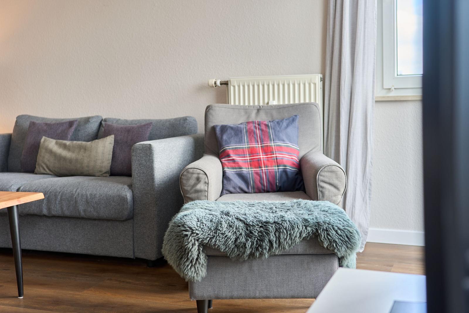 Ein grauer Sessel mit Kissen und Felldecke neben einem Sofa.