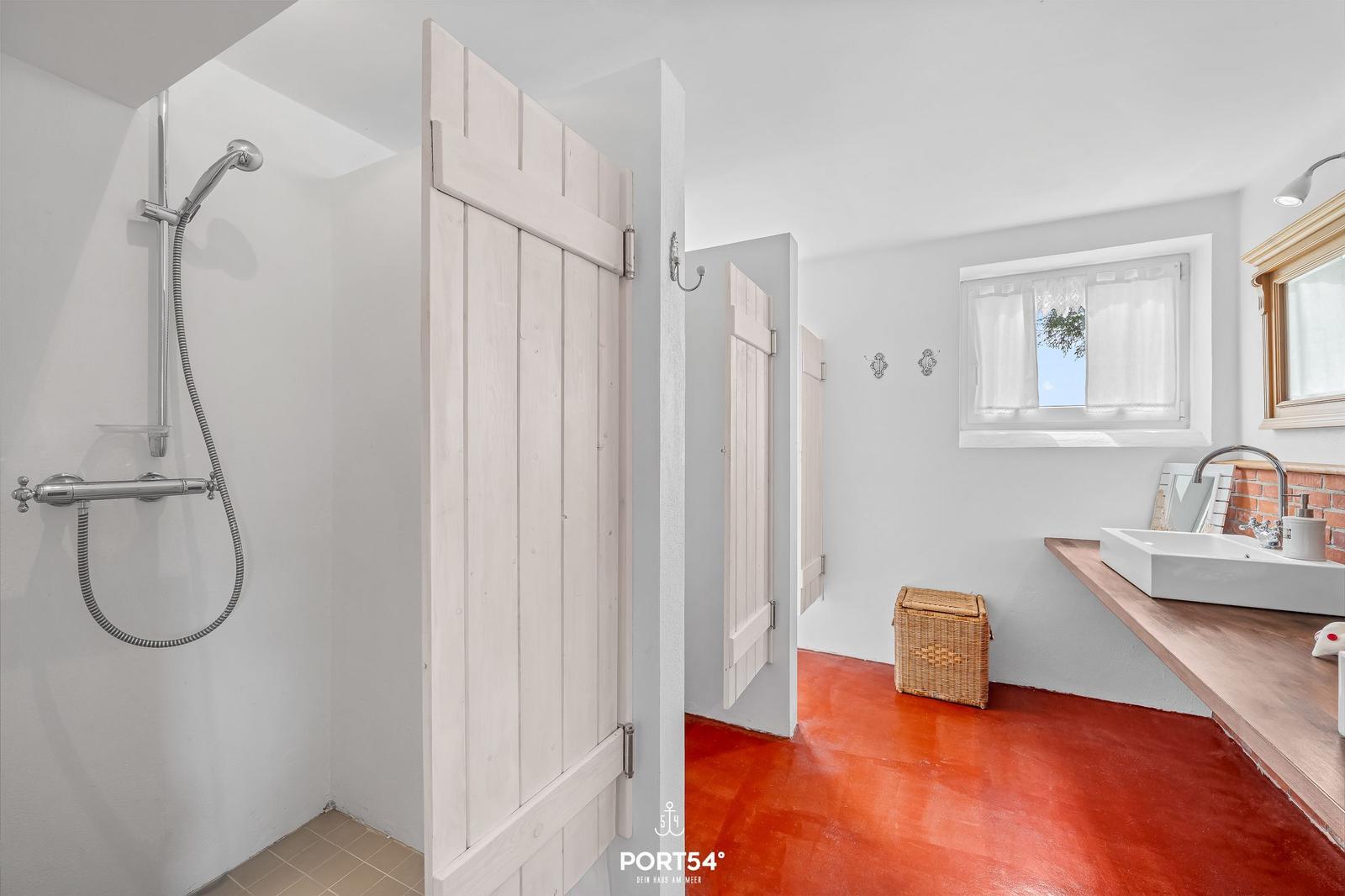 Bad mit Dusche, Waschbecken und rotem Boden. Holztür und Fenster mit Vorhängen.