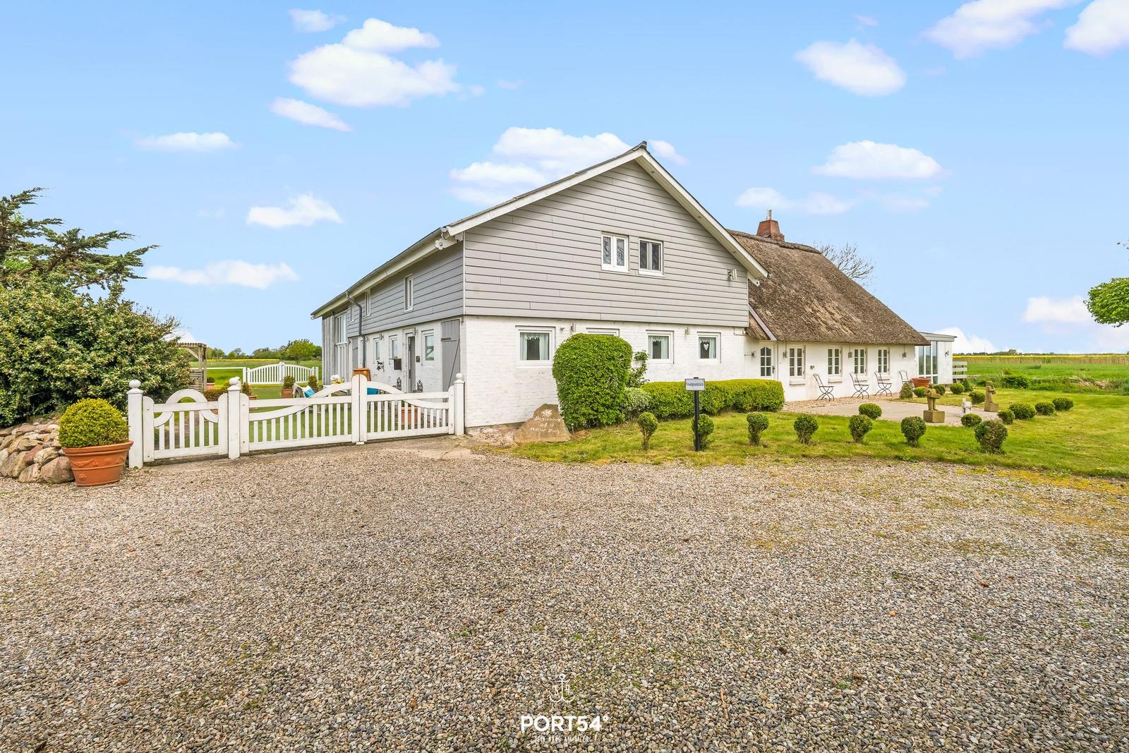 Haus mit Dach aus Stroh, weißer Putz und grauer Holzverkleidung. Vorgarten mit Gras, Sträuchern und weißem Zaun.