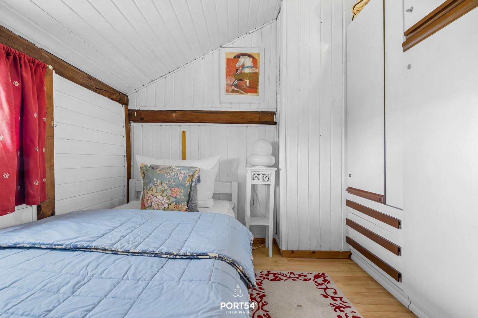 Schlafzimmer mit Bett, Nachttisch und Kleiderschrank. Weiße Wände, Holzbalken und rote Vorhänge.