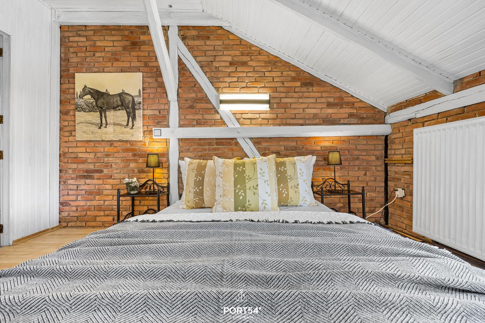 Schlafzimmer mit Bett, Kissen, Nachttischen und Backsteinwand unter schrägem Dach.