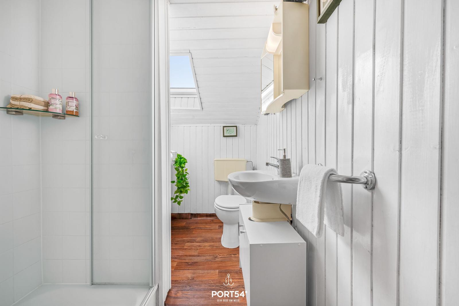 Badezimmer mit Dusche, Waschbecken und Toilette. Holzboden und weiß verkleidete Wände.
