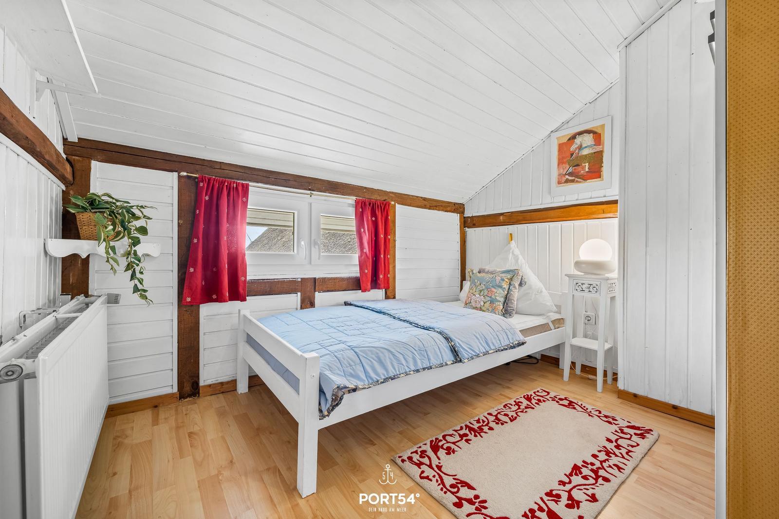 Schlafzimmer mit weißem Bett, Holzboden und roten Vorhängen an Fenstern.