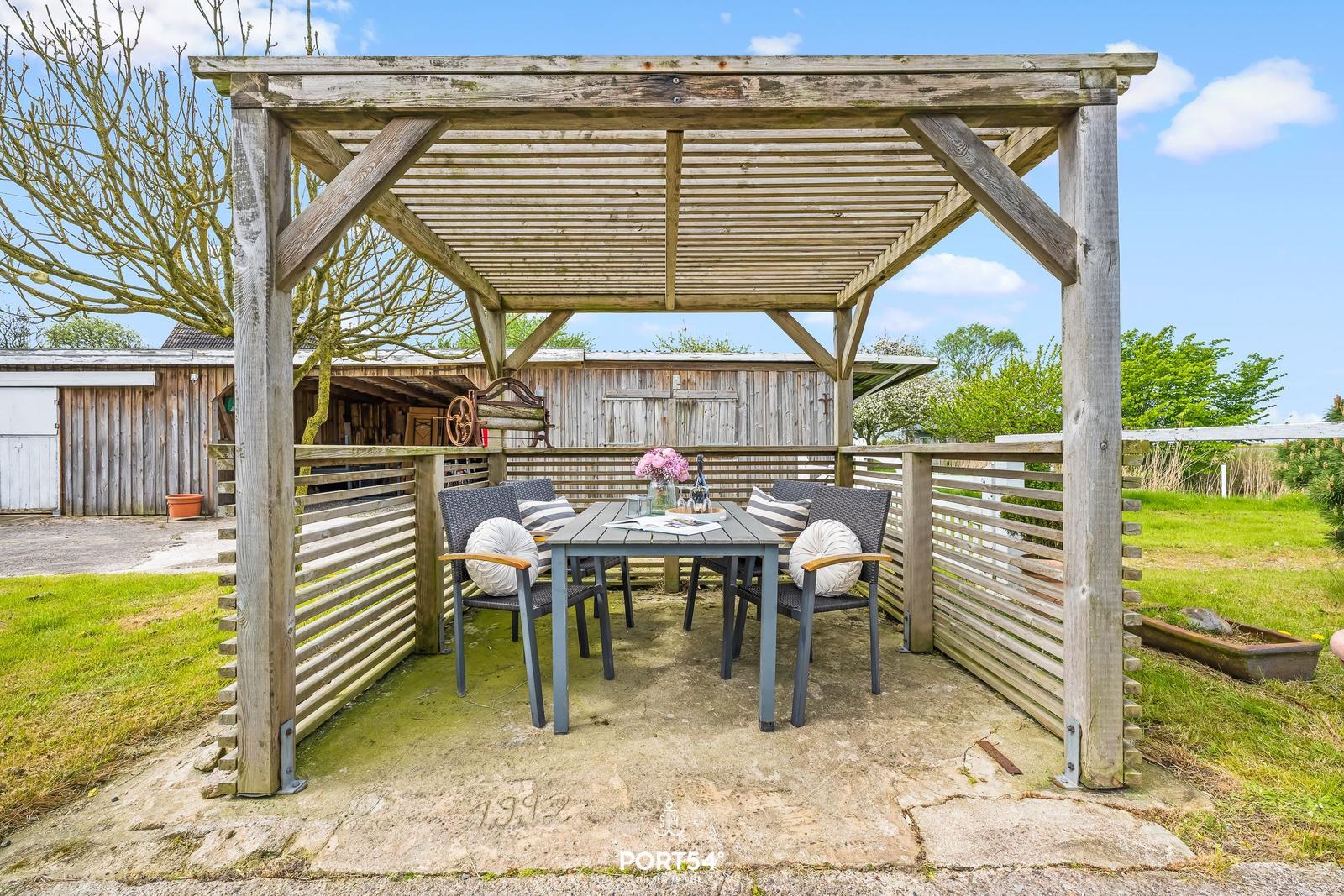 Holzüberdachter Terrassenbereich mit Tisch und Stühlen. Hintergrund: Holzgebäude und Grünfläche.