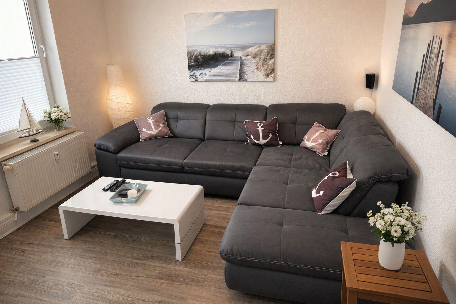 Wohnzimmer mit grauem Ecksofa, weißen Couchtisch und Seeblick-Bildern an der Wand.