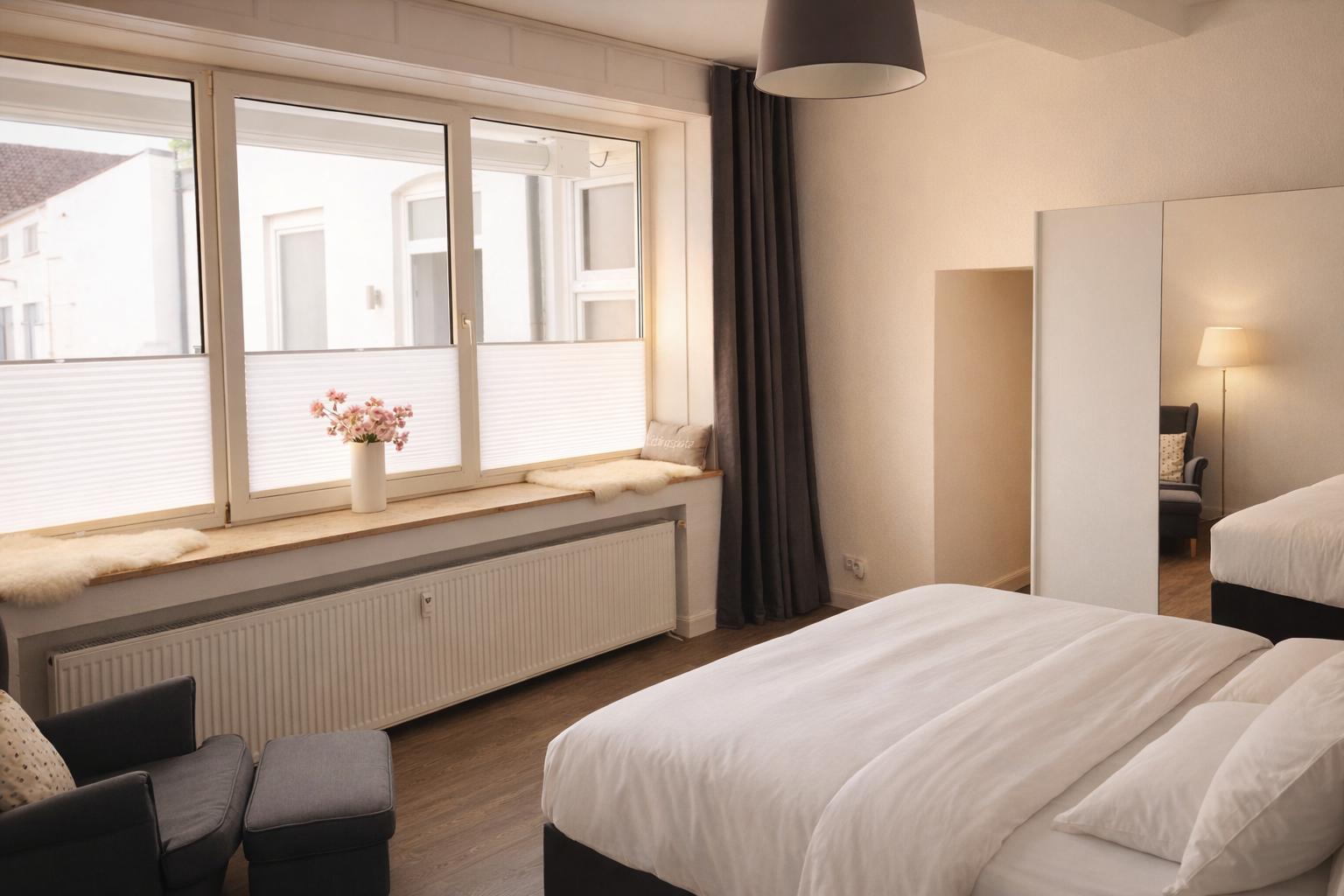 Schlafzimmer mit Bett, Fensterbank und Spiegel. Ein dunkler Sessel und eine Lampe sind sichtbar.