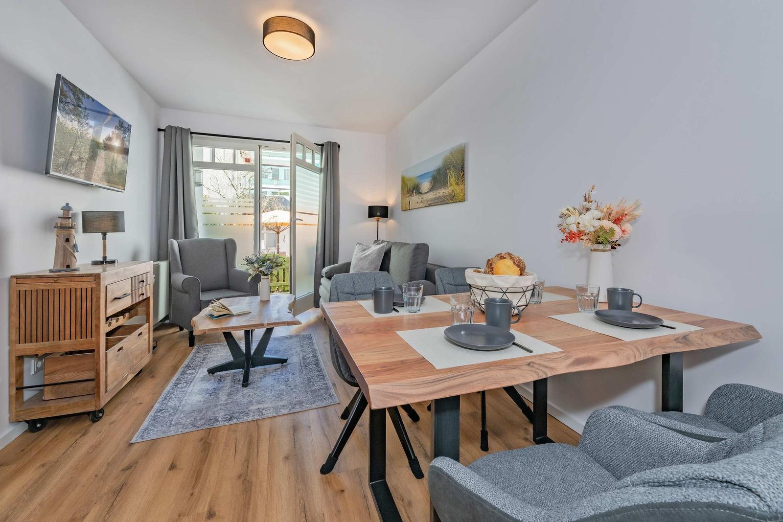 Wohn- und Essbereich mit Holztisch, grauen Sitzgelegenheiten und Fernseher an der Wand.