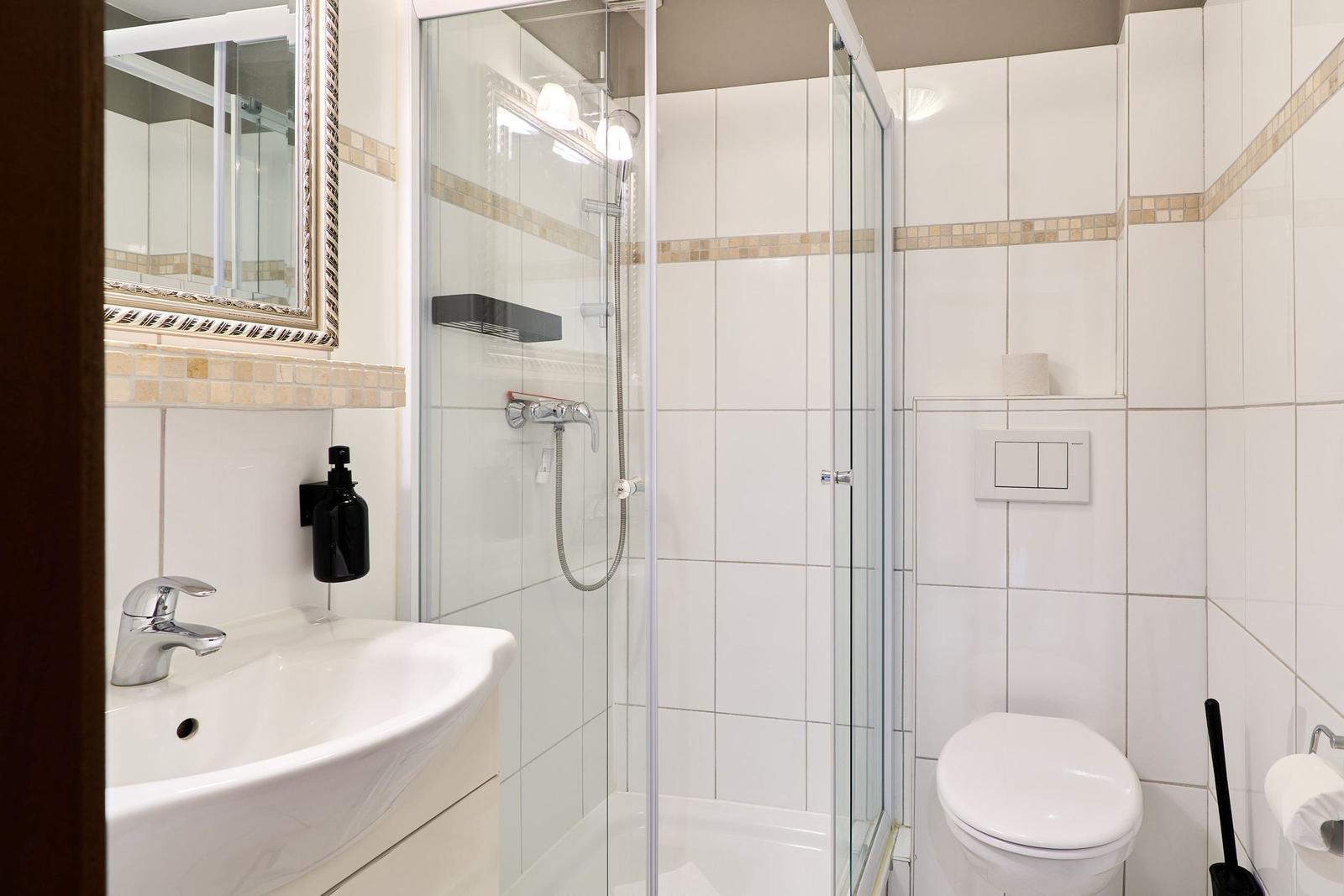Badezimmer mit Dusche, Waschbecken und Toilette. Weiße Fliesen und Spiegel.