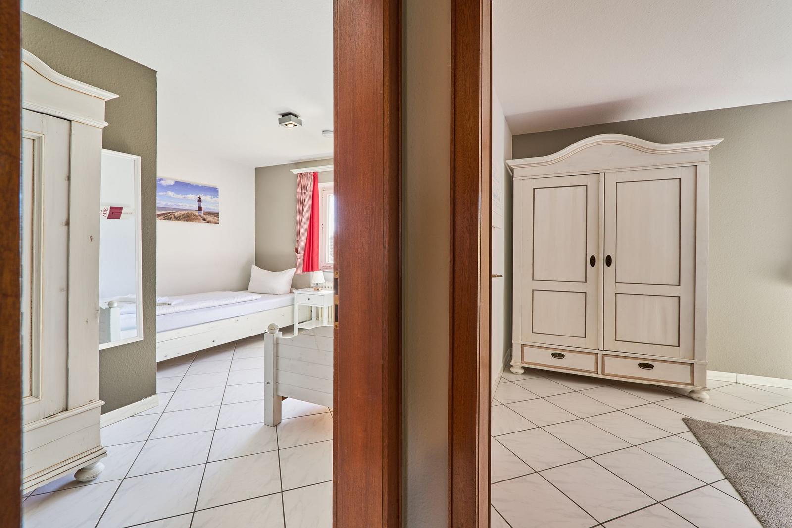 Schlafzimmer mit weißem Bett und Kleiderschrank. Blick durch Tür auf weitere Räume.