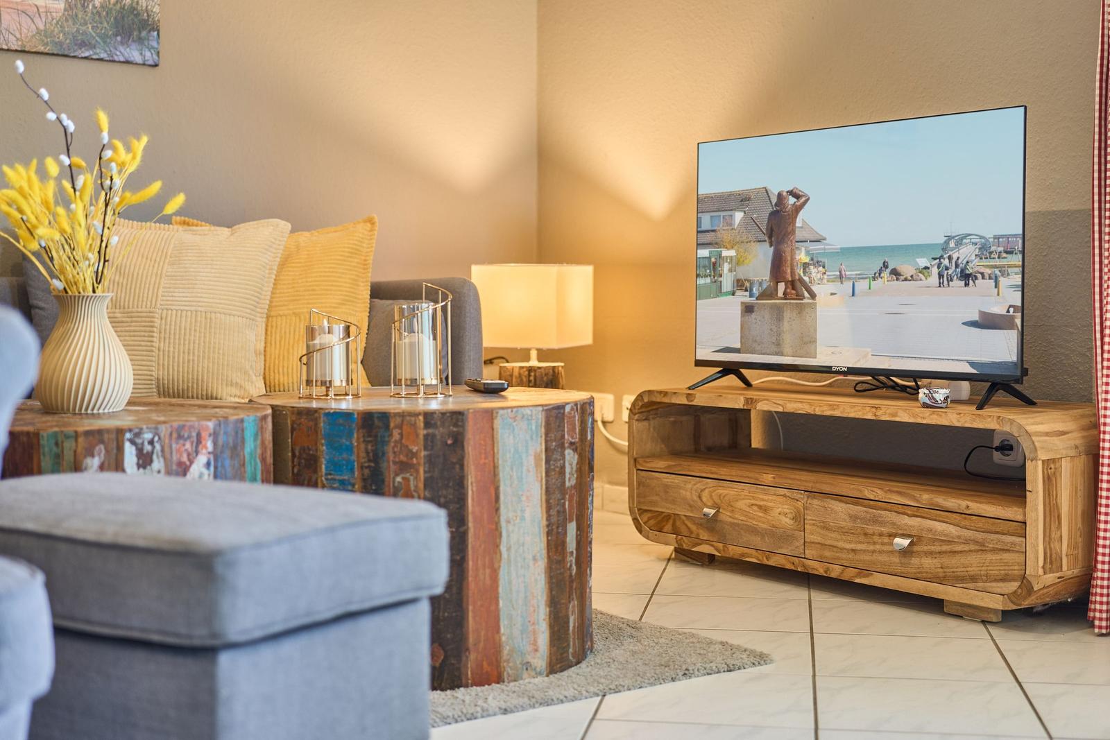 Ein Wohnzimmer mit Sofa, TV und Holztisch. Auf dem Bildschirm ist eine Küstenansicht zu sehen.